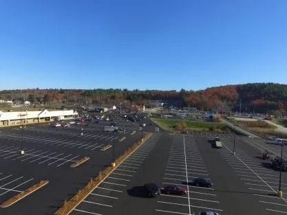 An aerial view of a large, mostly empty asphalt parking lot with a building on the left under a clear blue sky.