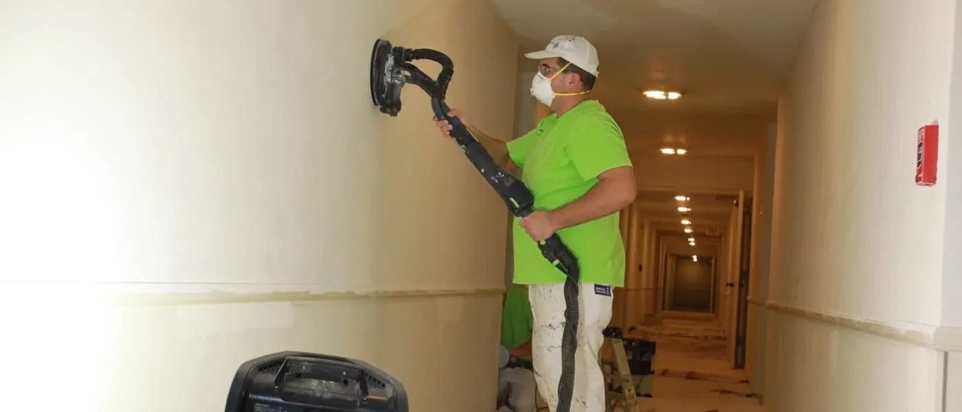 A worker in a bright green shirt uses a long-handled sander on a wall in a hallway.