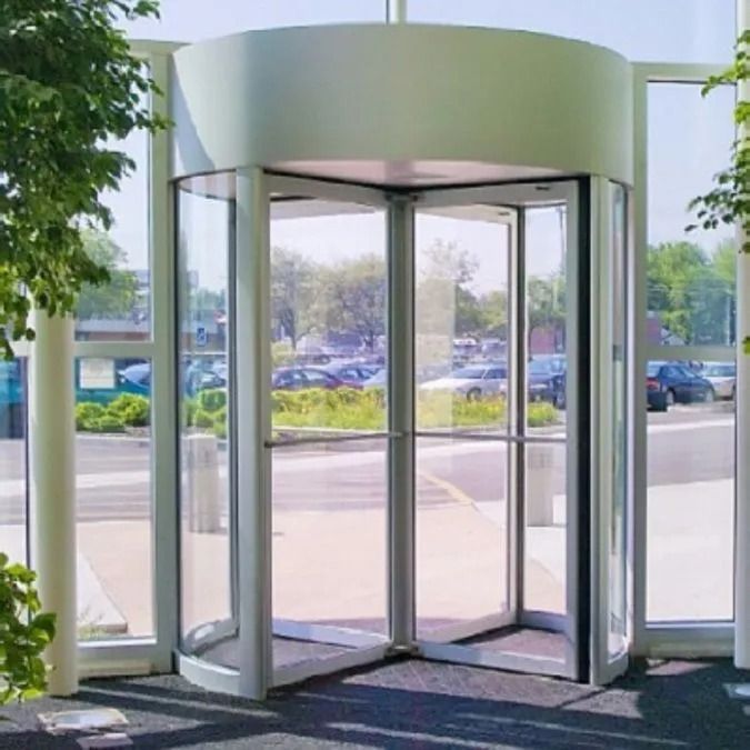 A glass revolving door at an entrance with a white frame and cylindrical top, looking out onto a parking lot.