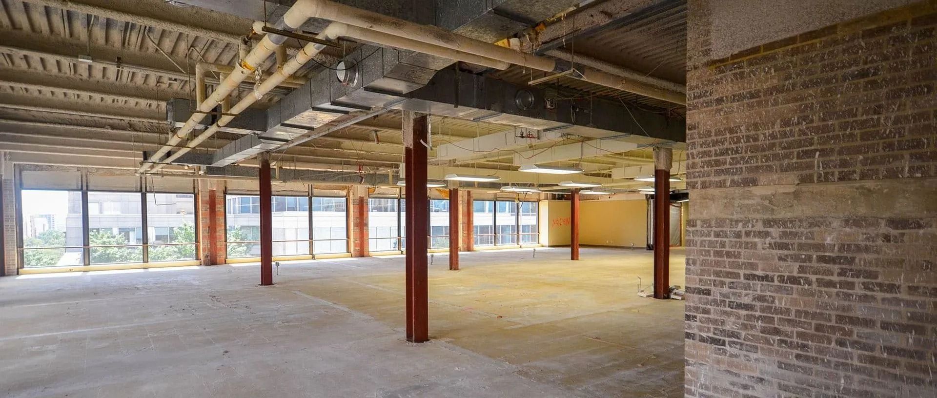 An unfinished, open-plan office space with exposed ceiling pipes, concrete floors, red steel columns, and brick walls.