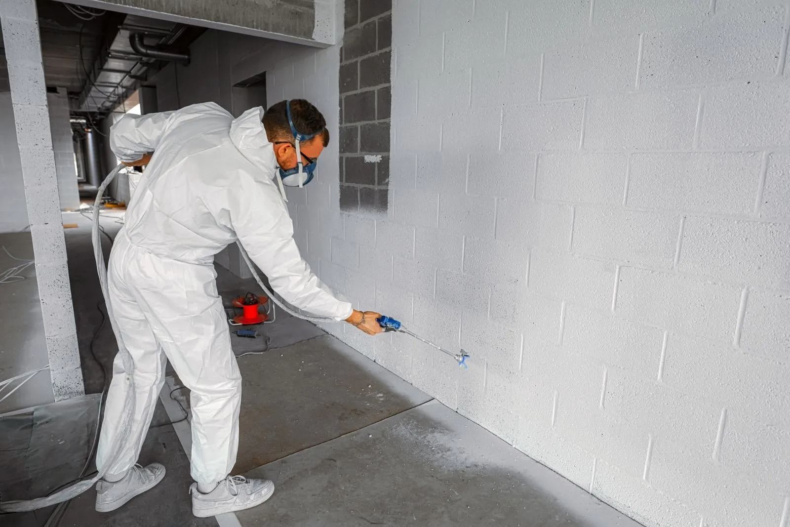 A person in white protective gear and a face mask uses a spray gun to paint a white concrete wall in an indoor space.