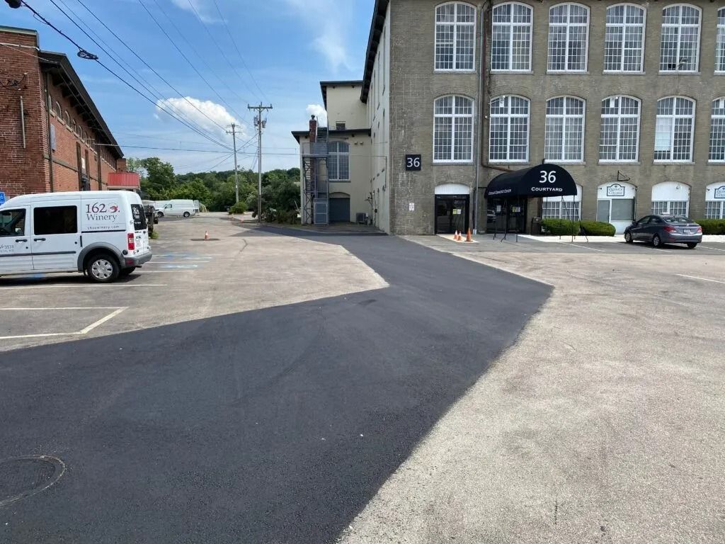A parking lot with new black asphalt patches in front of a brick building with a dark awning labeled 