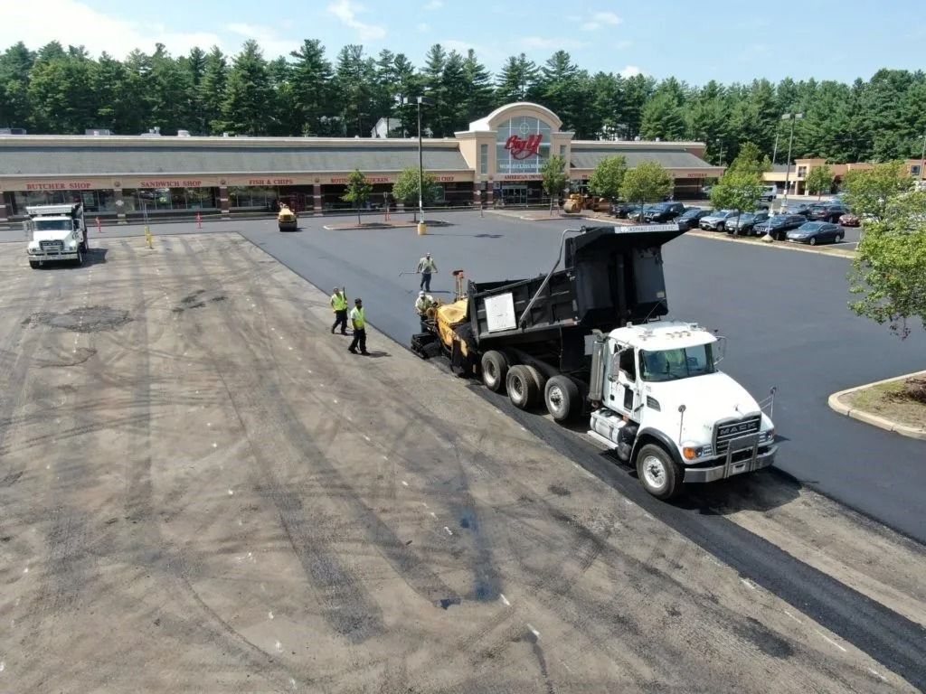 A construction crew uses a dump truck and paving equipment to lay fresh asphalt on a parking lot at a shopping center.