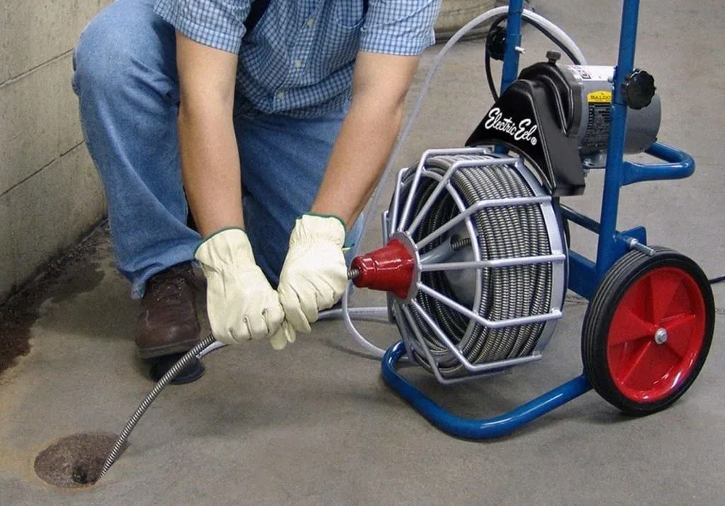 A person wearing protective gloves feeds a metal drain cleaning cable into a floor drain using a portable electric machine.