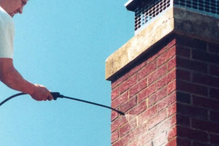 A person uses a pressure washer to clean a red brick chimney outdoors against a clear blue sky.