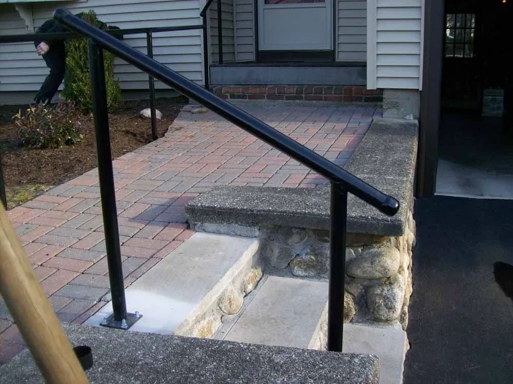 A black metal handrail installed on a stone step leading to a brick walkway and a house entrance.