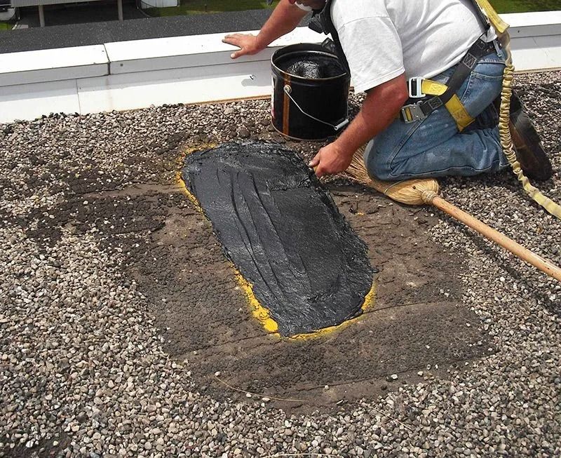 A worker in safety gear spreads black roofing sealant over a gravel roof patch.