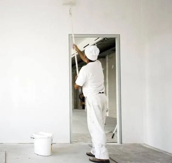 A person wearing white professional clothing uses a long-handled paint roller to paint a wall white.
