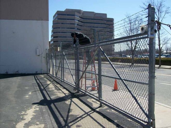 Person climbing a chain-link fence beside a road, with orange cones and a concrete building nearby.