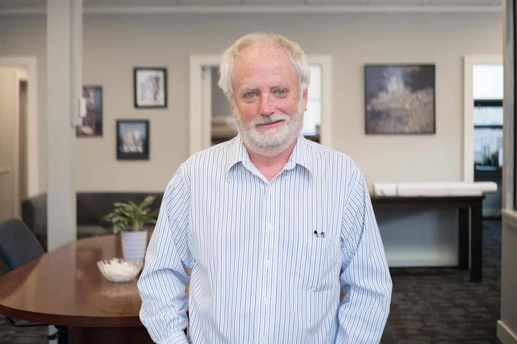 Man standing in a modern office lobby, wearing a light striped shirt and smiling at the camera.
