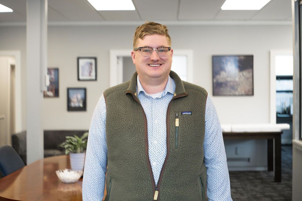 Man standing in an office, smiling in a green vest and patterned shirt.