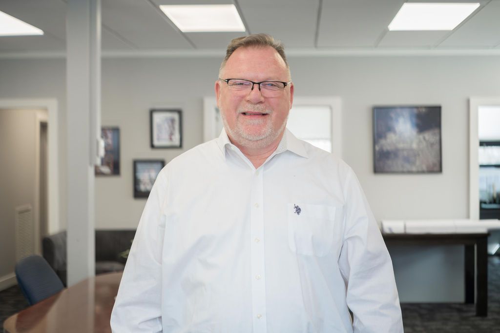 Man standing in a bright office, wearing a white shirt and smiling at the camera.