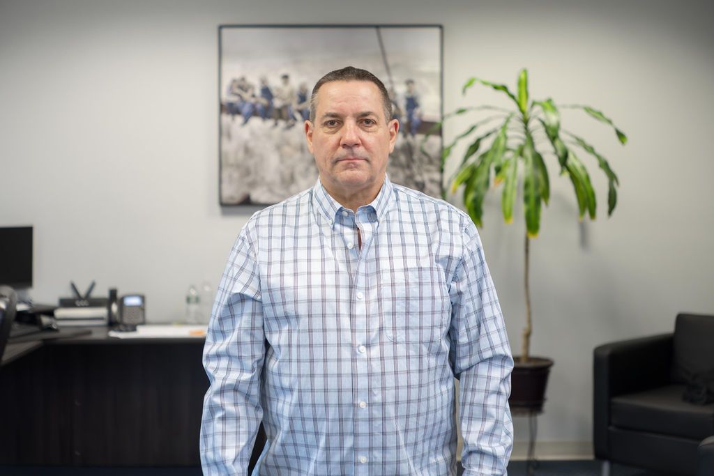 Man standing in an office, wearing a blue checkered shirt, with a plant and wall art behind him.
