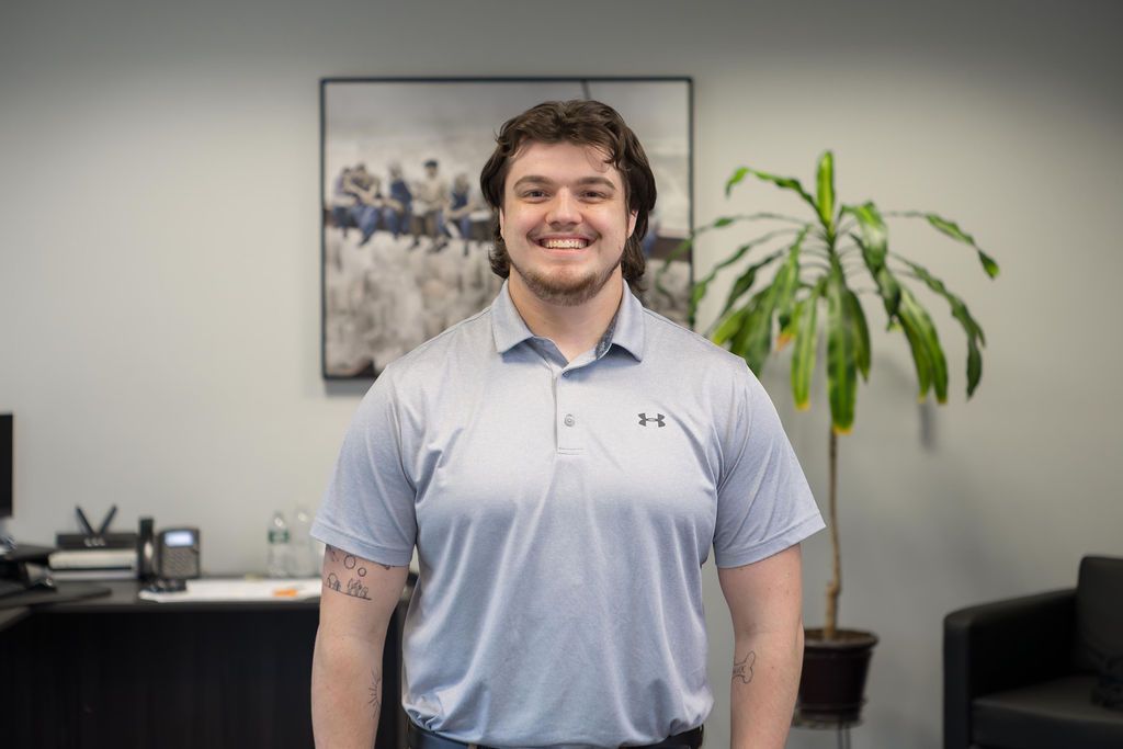 Smiling person in a light blue polo standing in an office with a plant and framed wall art.