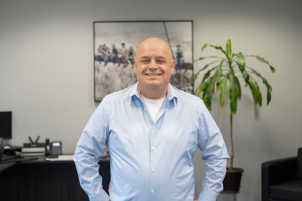 Man standing in an office, wearing a light blue checkered shirt, with a plant and wall art behind him.