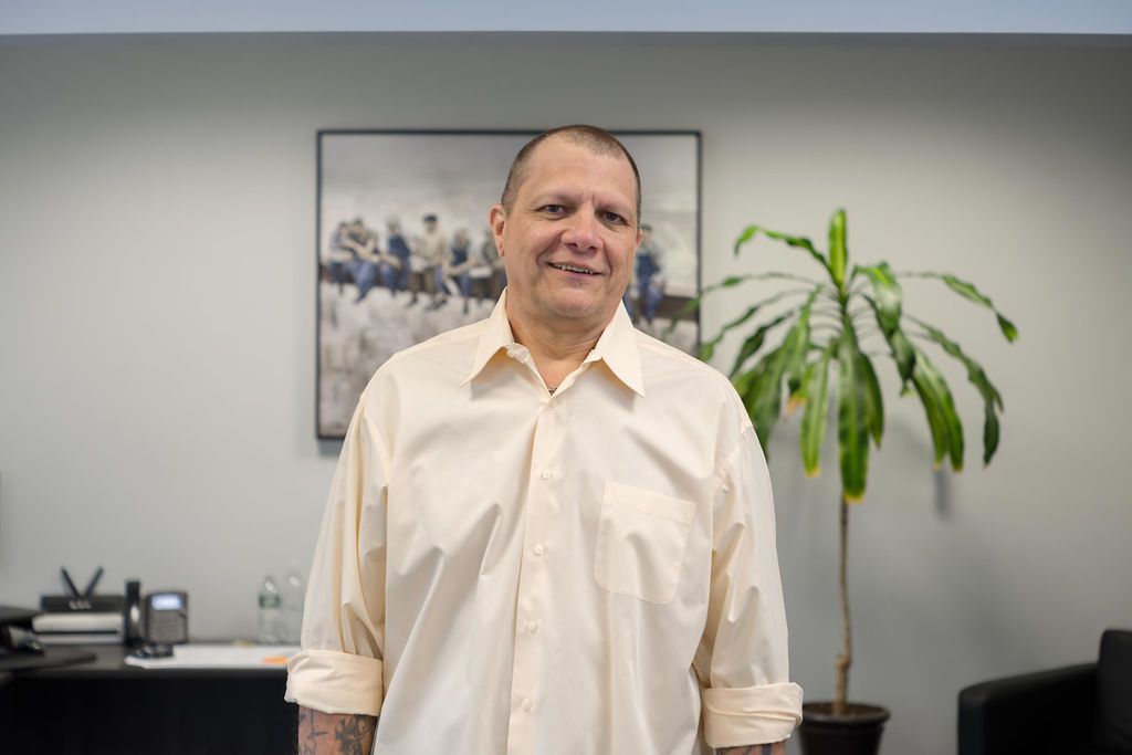 Man in a white shirt standing in an office with a plant and framed wall art behind him