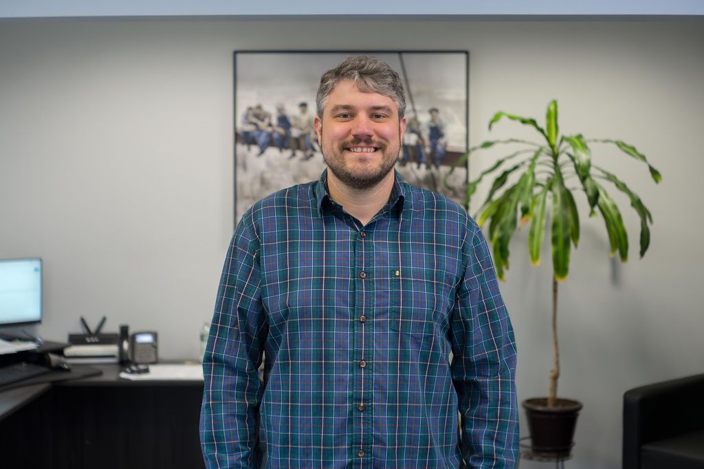 Man standing in an office, wearing a blue plaid shirt, smiling with a plant and framed art behind him.