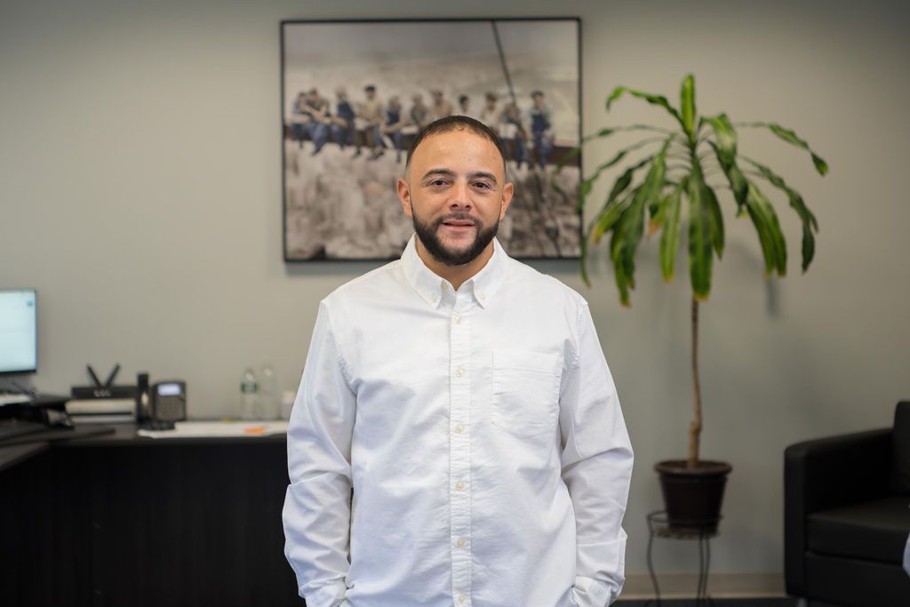 Man in a white shirt standing in an office with a desk, plant, and framed wall art behind him