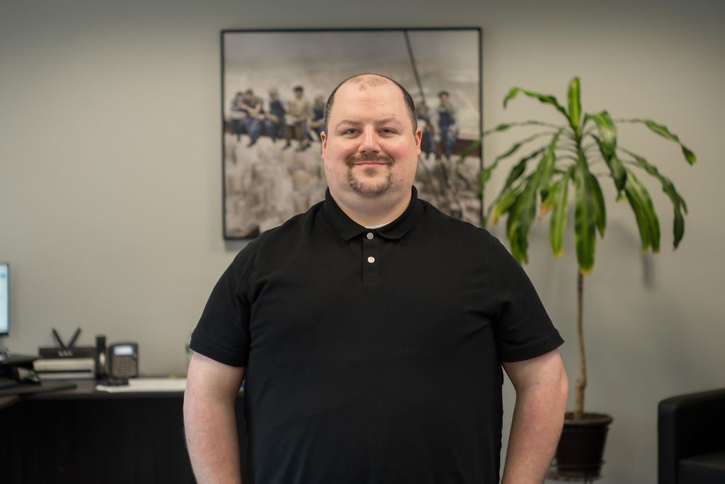 Man standing indoors in a black polo, smiling in an office with a plant and wall art behind him.