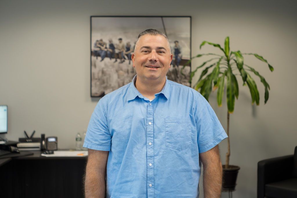 Smiling man in a light blue shirt stands in an office with a plant and framed wall art behind him