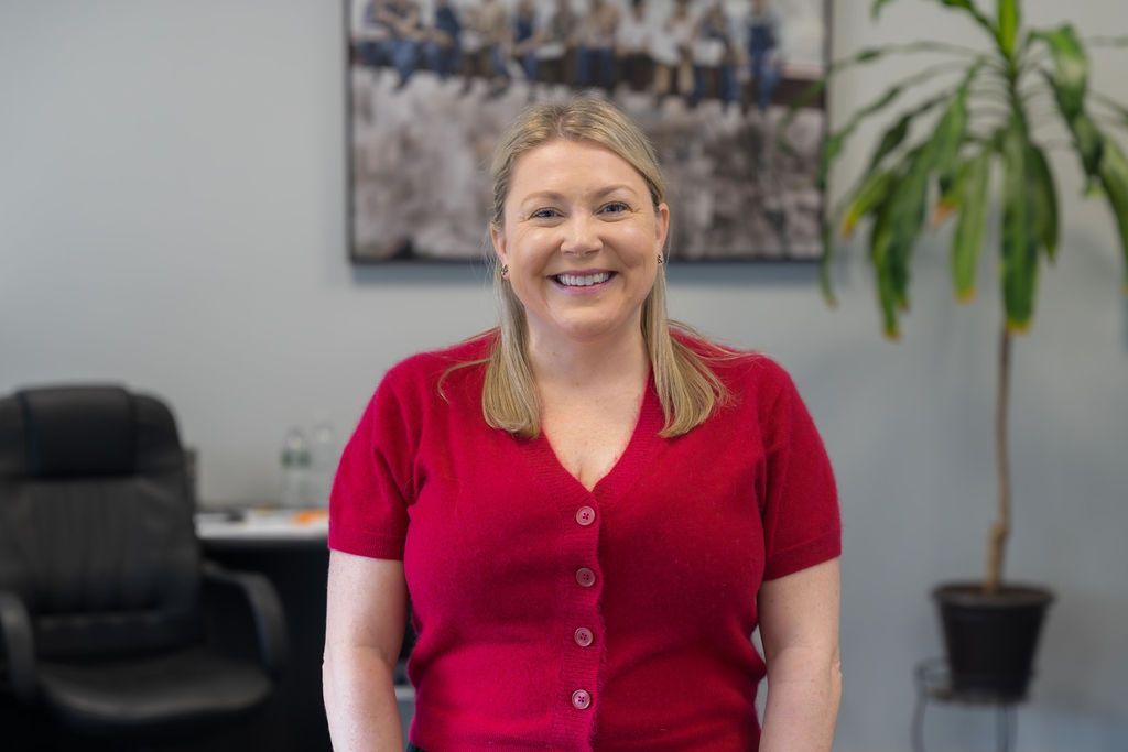 Smiling woman in a red cardigan standing in an office with a chair, wall art, and a potted plant.