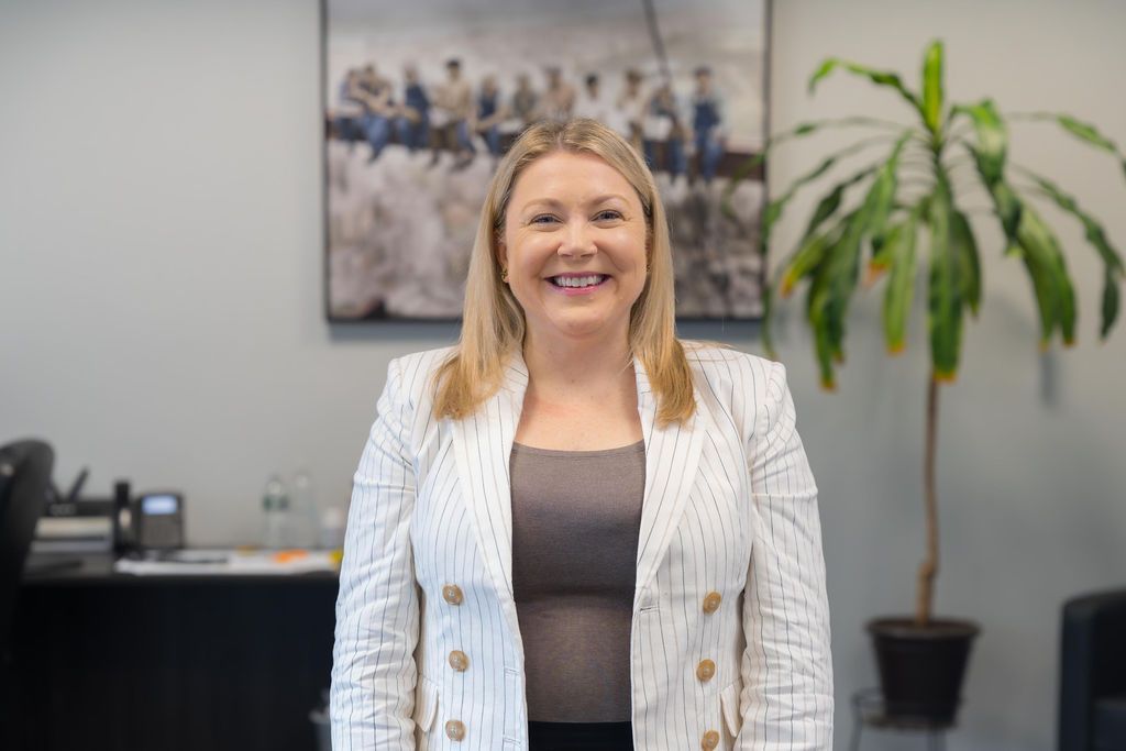 Woman standing in an office, smiling in a white blazer with a plant and desk behind her