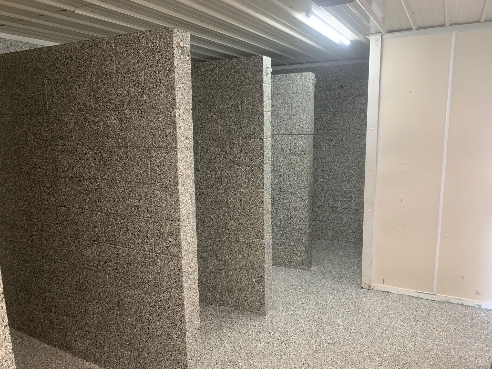 Small room with speckled walls and floor, divided by narrow partitions and lit by a corrugated ceiling.