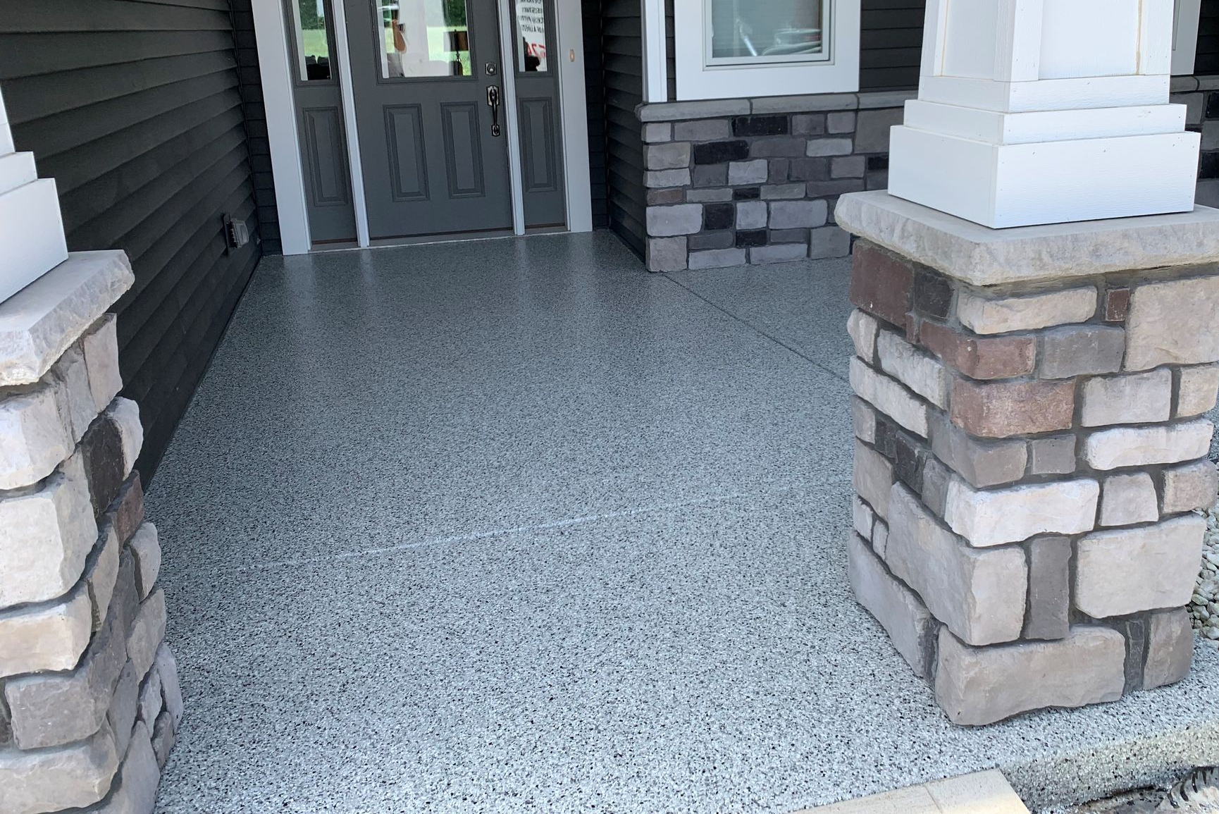 Front porch with gray speckled floor, dark double doors, and stone columns beside a house entrance