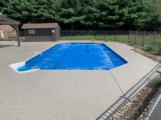 Covered outdoor swimming pool with a blue winter cover and concrete deck surrounded by fencing and trees