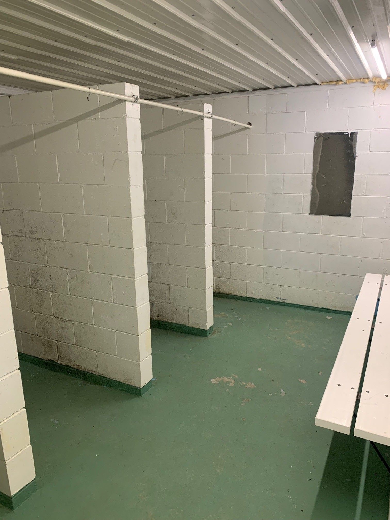 Empty restroom with white stall walls, green floor, and a bench along the right wall.