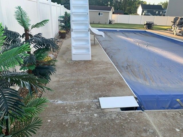 Backyard pool with white slide, blue cover, and concrete patio beside a fence