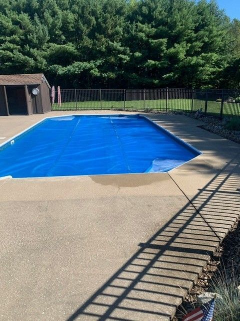 Covered outdoor swimming pool beside a tan deck, with a black fence and trees in the background.