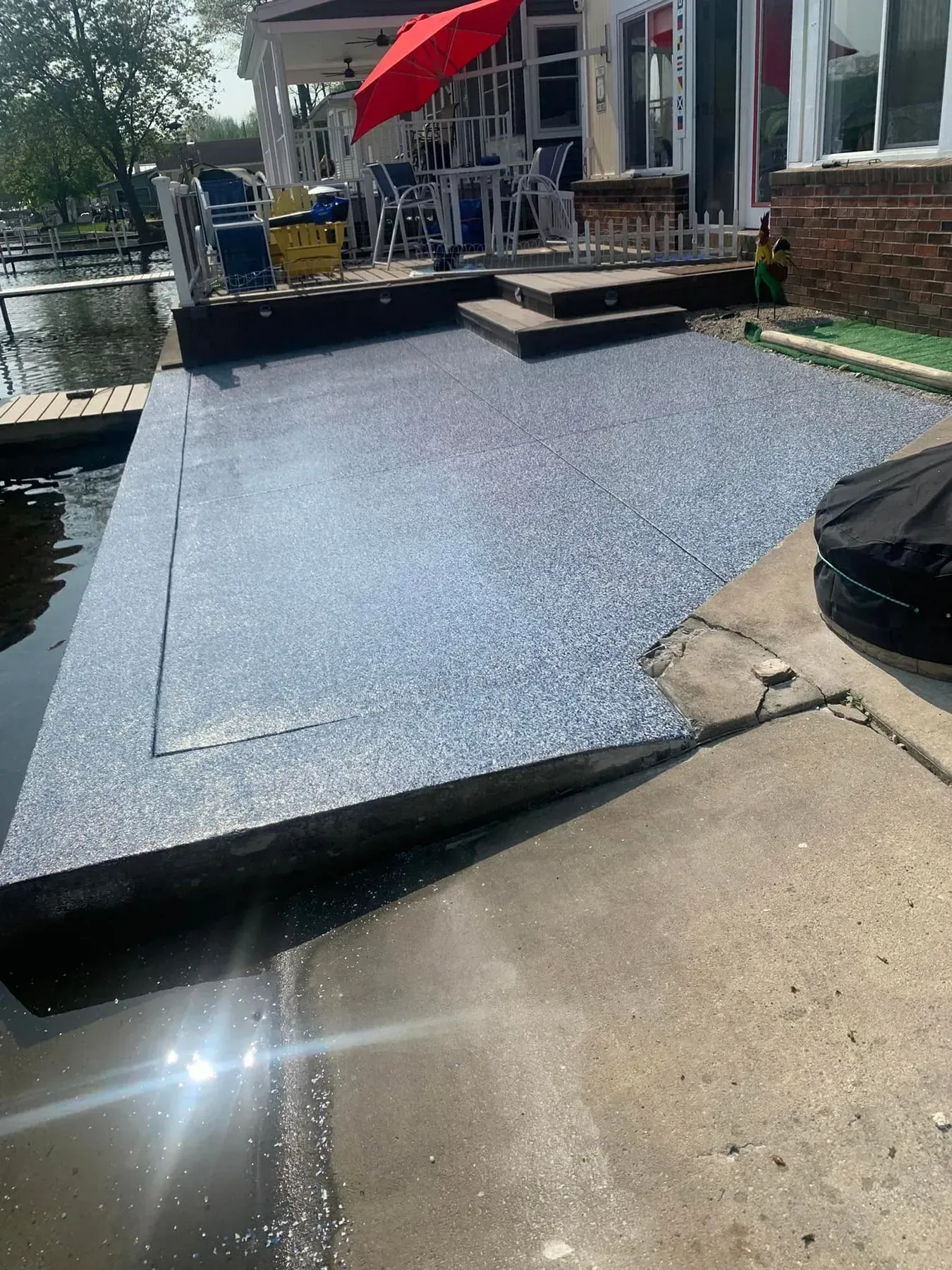 Sloped dark-blue dock ramp beside a waterfront patio with a red umbrella and house exterior