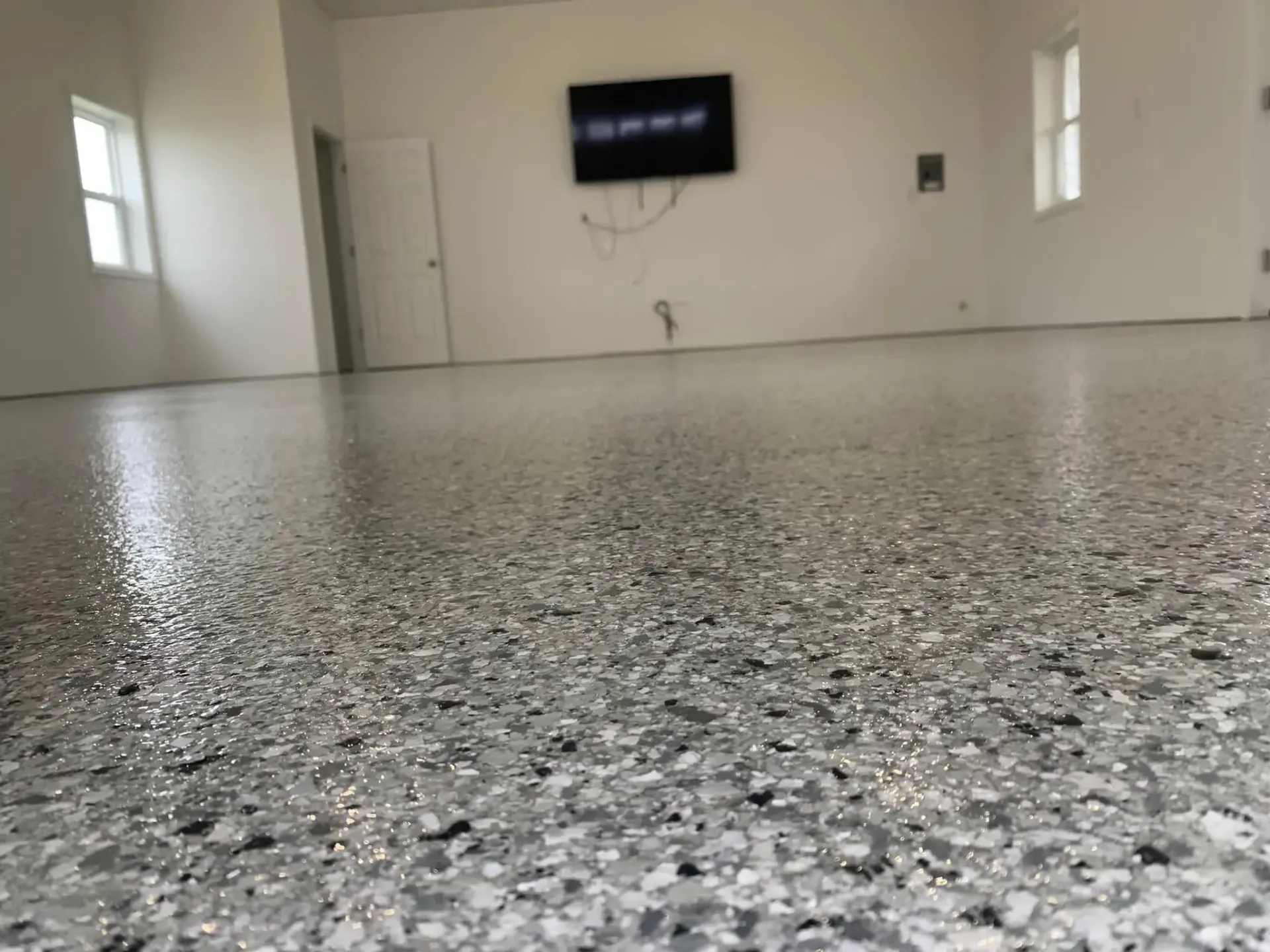 Empty room with speckled gray floor, white walls, small windows, and a wall-mounted TV.