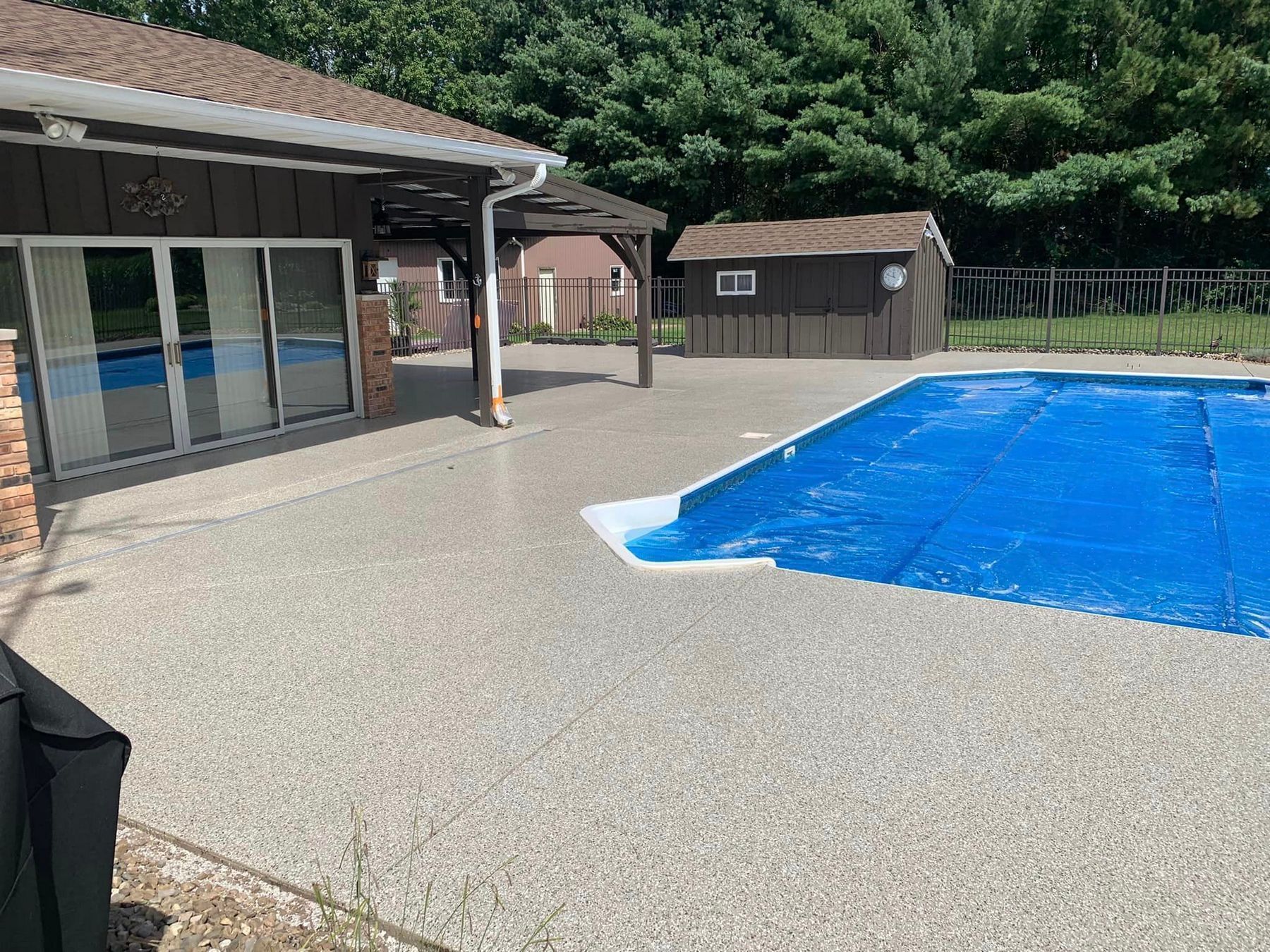 Backyard pool beside a house with patio, blue cover, and wooded hillside in the background