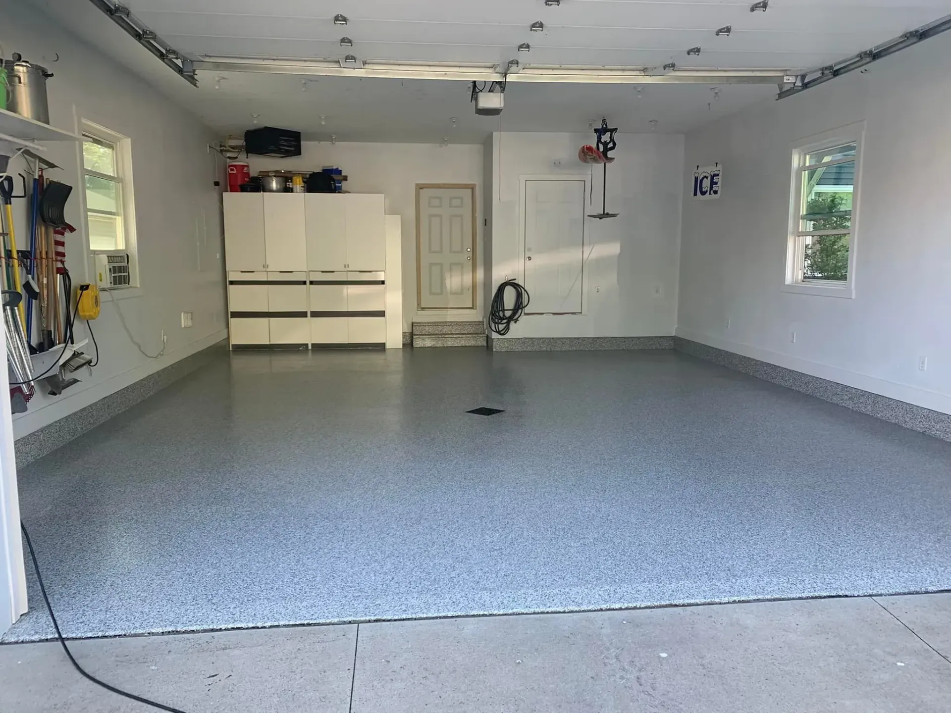 Empty garage with gray speckled floor, white walls, cabinets, and windows.