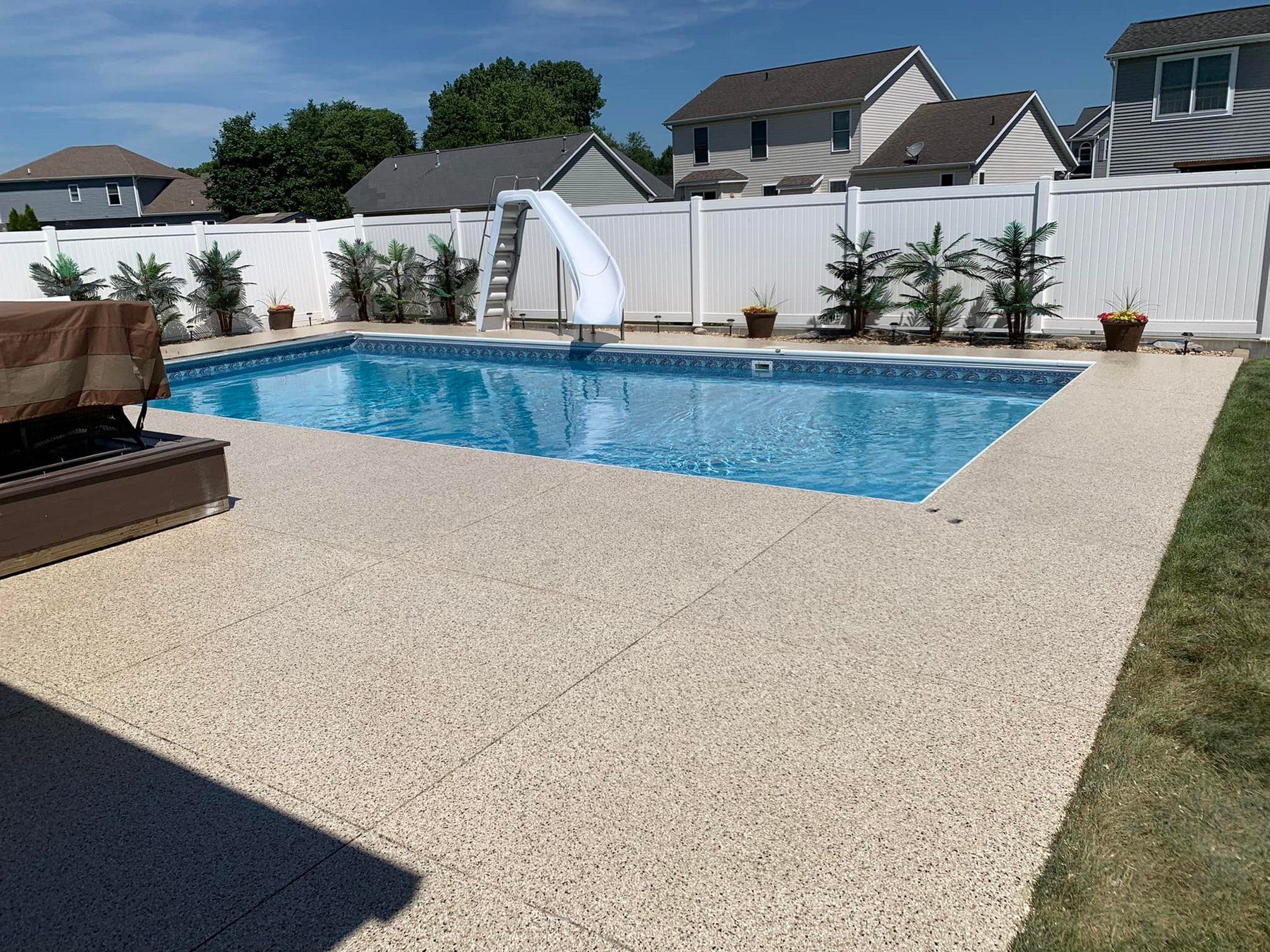 Backyard pool with blue water, white slide, and stone patio beside a white fence and suburban homes