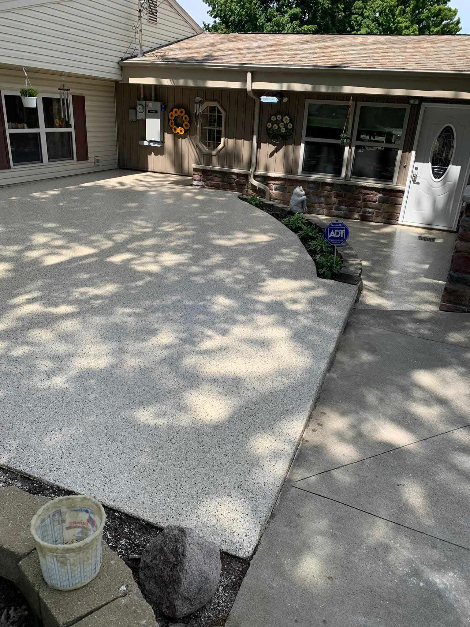 Freshly poured concrete patio beside a house, with a bucket in the foreground and shaded walkway nearby