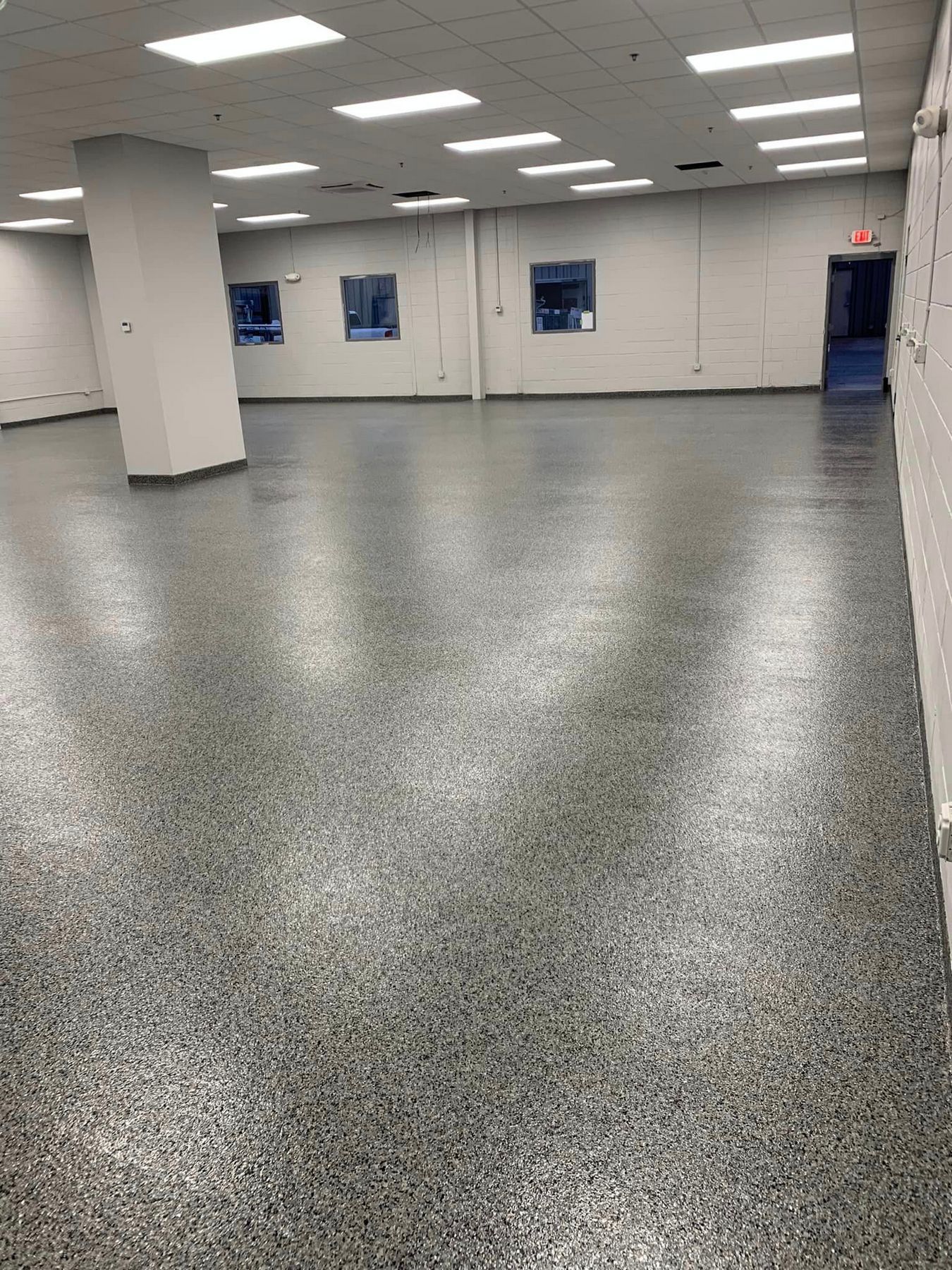 Empty fluorescent-lit room with gray speckled floor, white walls, and a central support column