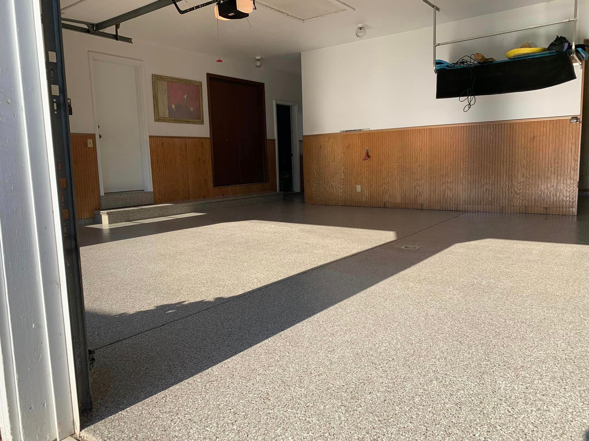 Empty garage with speckled floor, wood-paneled walls, and sunlight streaming through the open door