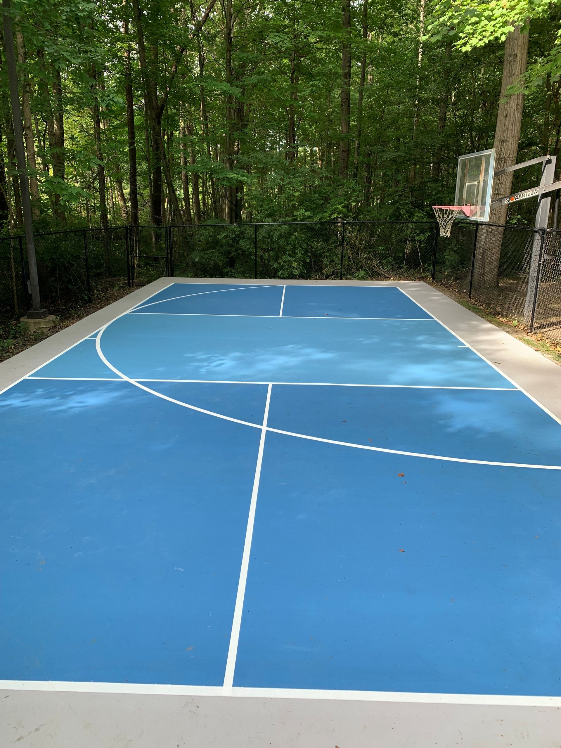 Outdoor blue basketball court in a wooded area, with white lines and a hoop on the right.