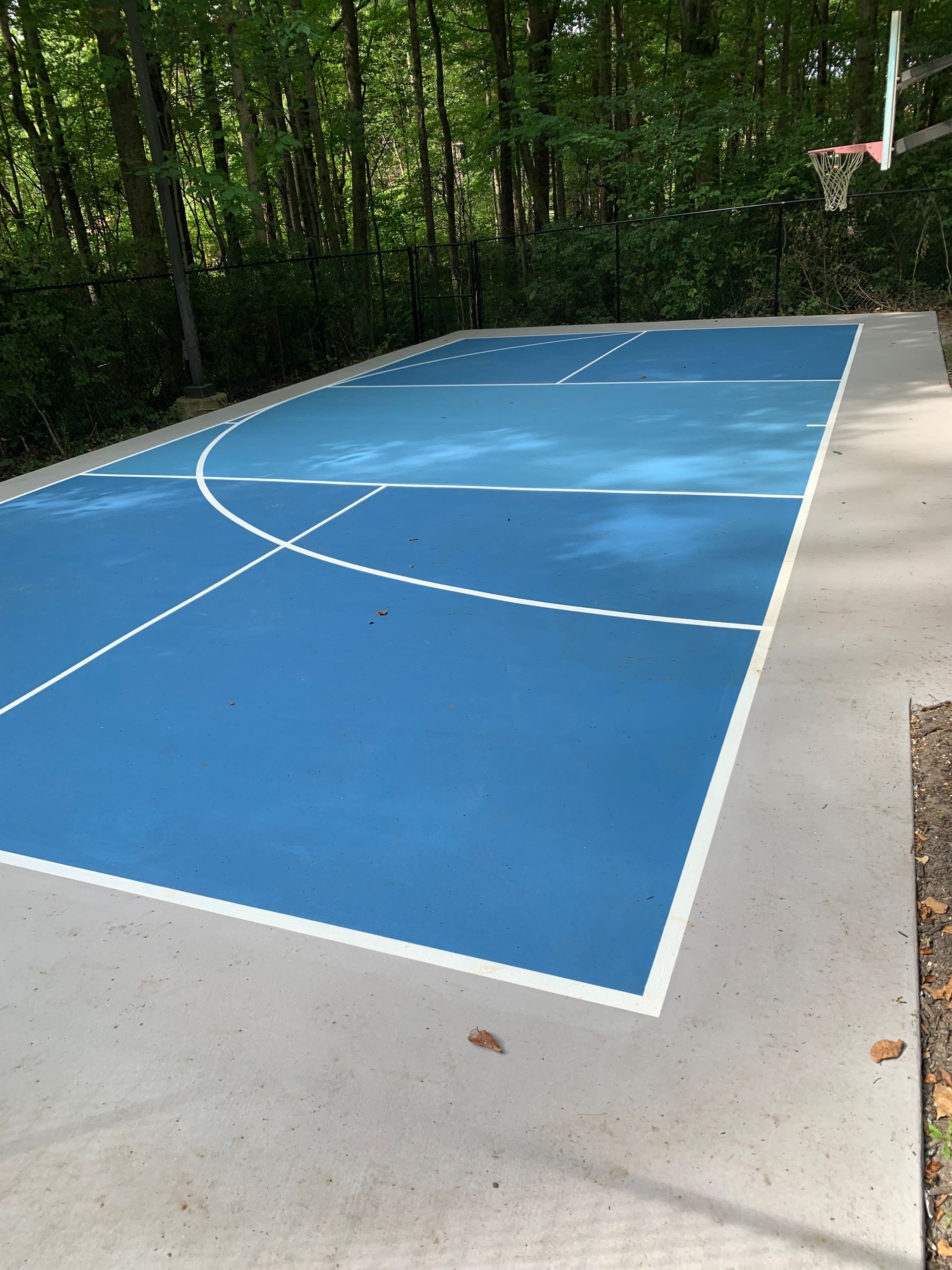 Blue outdoor basketball court surrounded by trees, with a hoop on the right side.