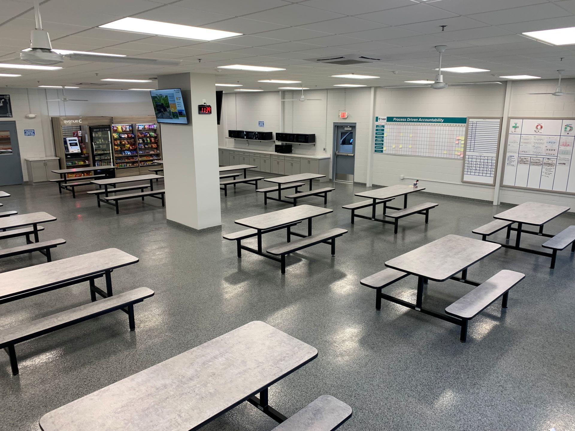 Empty school cafeteria with rows of gray tables and benches under bright fluorescent lights.