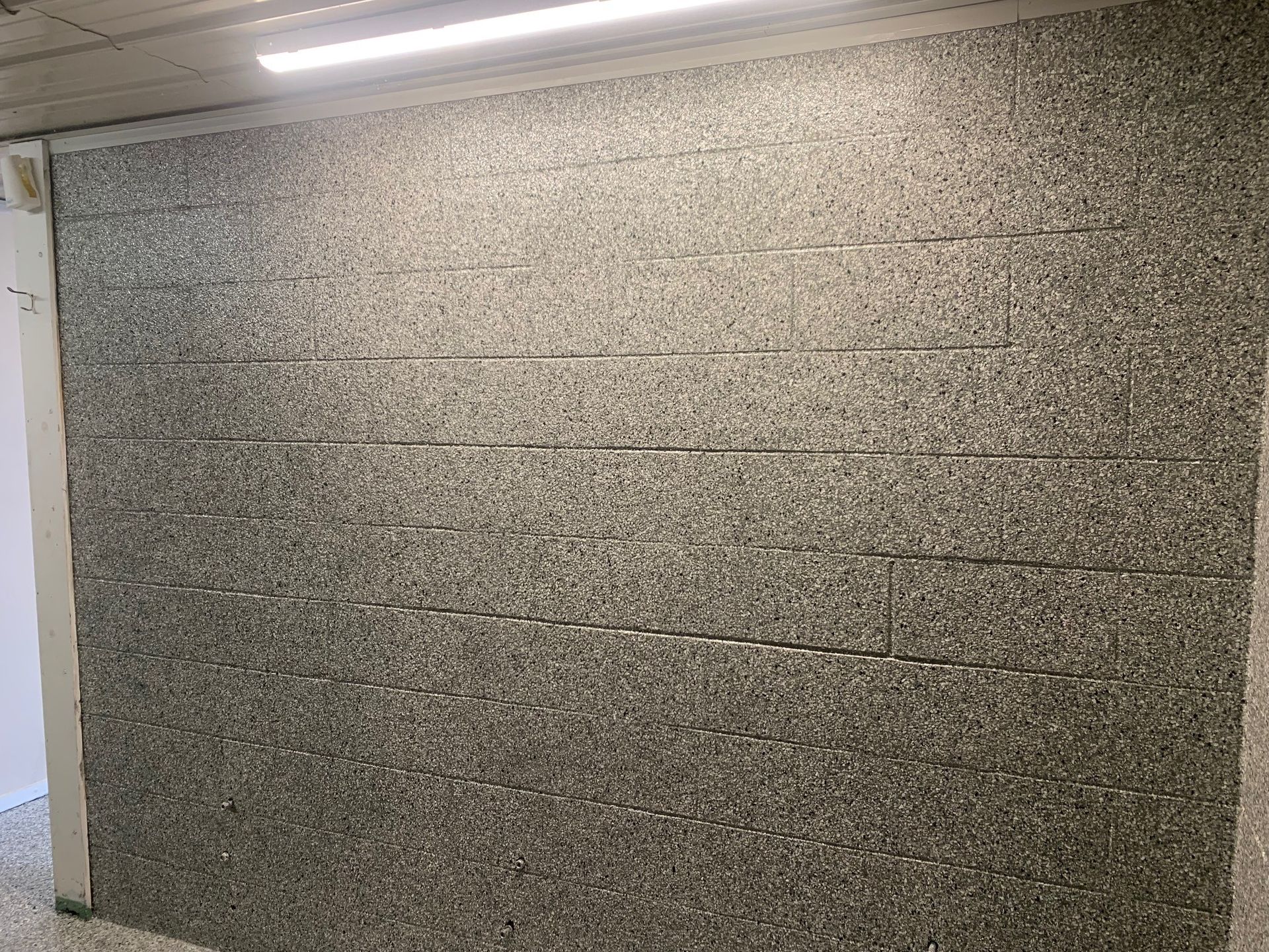 Gray tiled wall in an indoor hallway with fluorescent lighting and a doorway on the left.