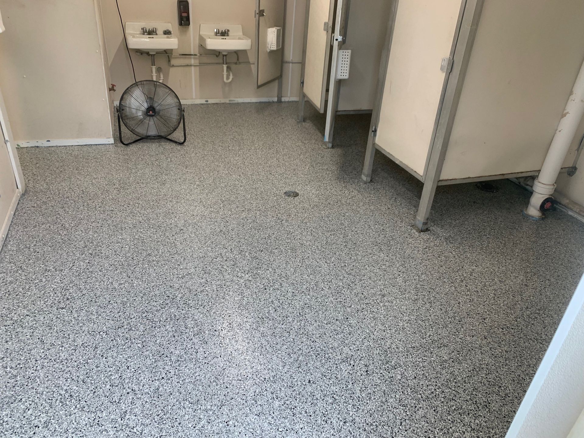 Empty restroom stall area with gray speckled floor, sink, and floor fan near the wall