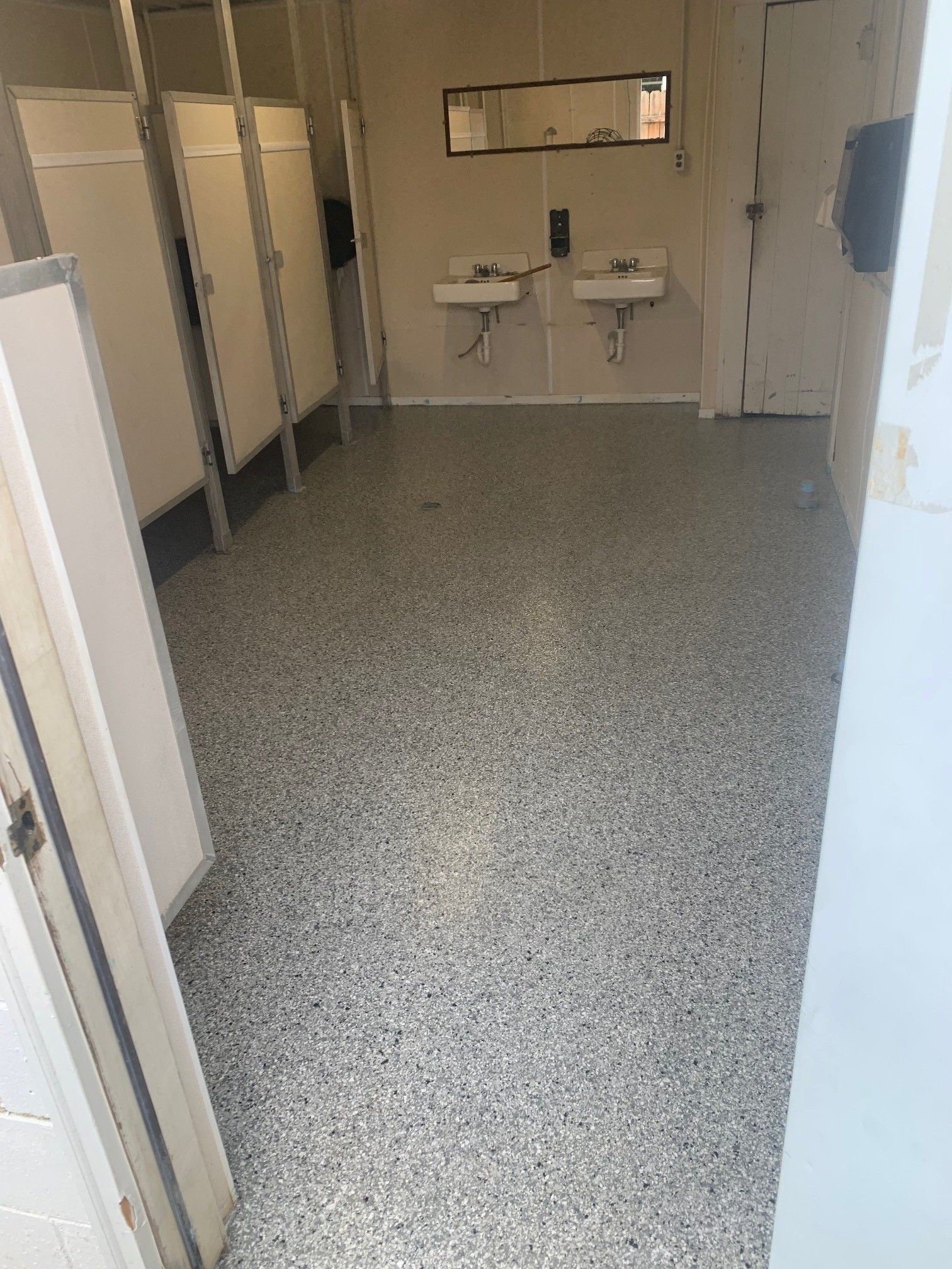 Empty restroom with speckled floor, stalls on the left, and two sinks on the back wall.