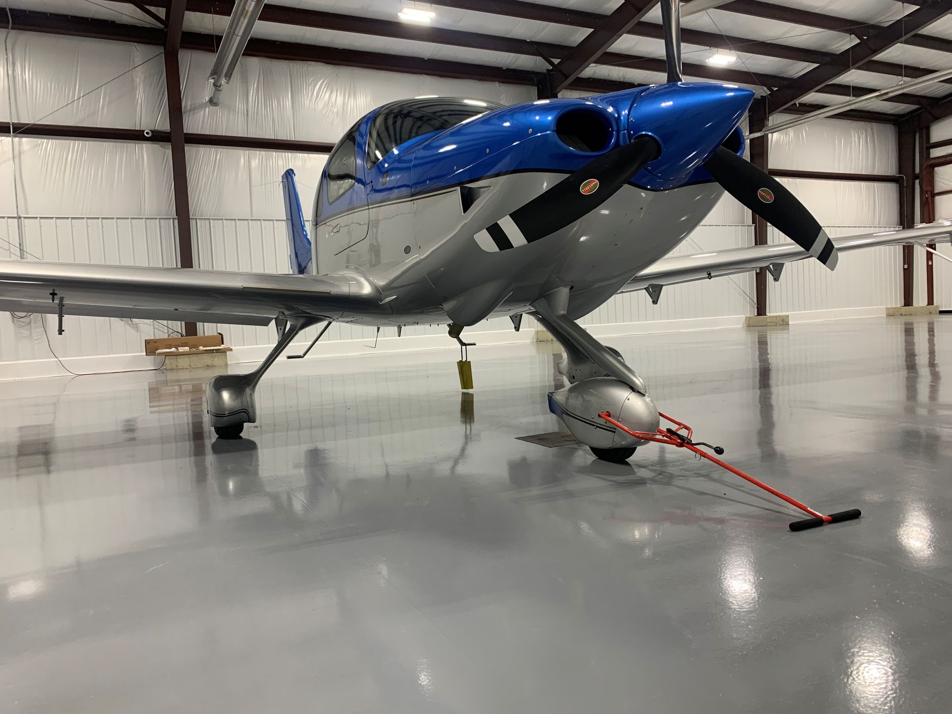 Blue single-engine airplane in a hangar, secured with a red tie-down strap on a polished floor.