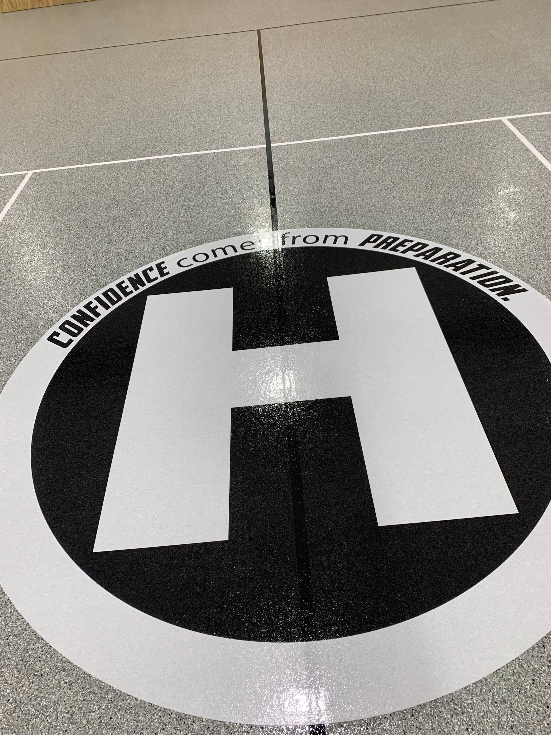 Helipad marking with a large white H inside a black circle on concrete pavement.