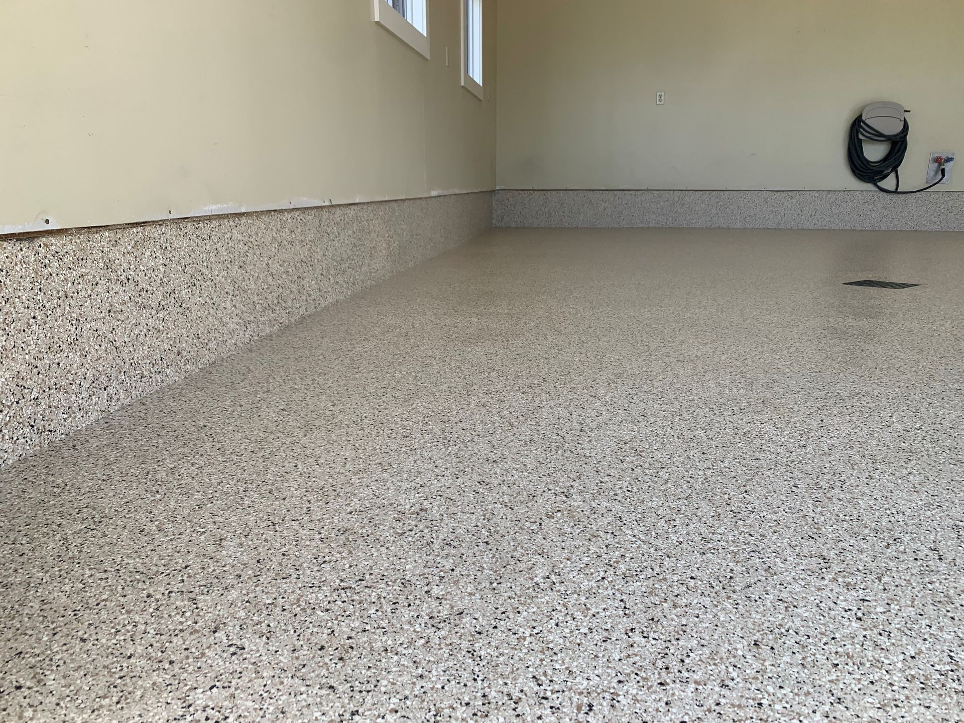 Empty indoor room with speckled floor, beige walls, and a wall outlet on the right