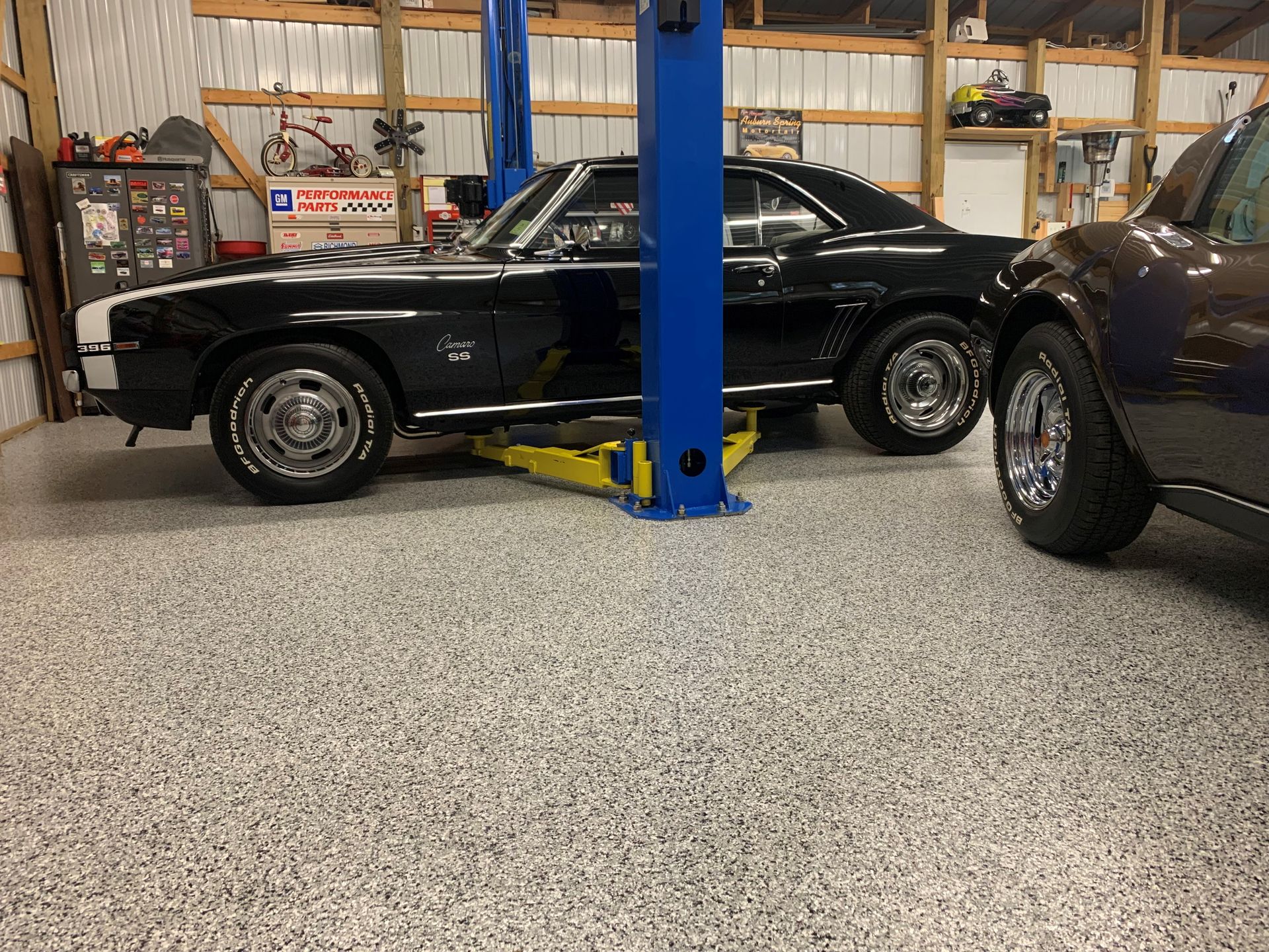Black classic muscle car in a garage lift bay, with another car partially visible beside it.